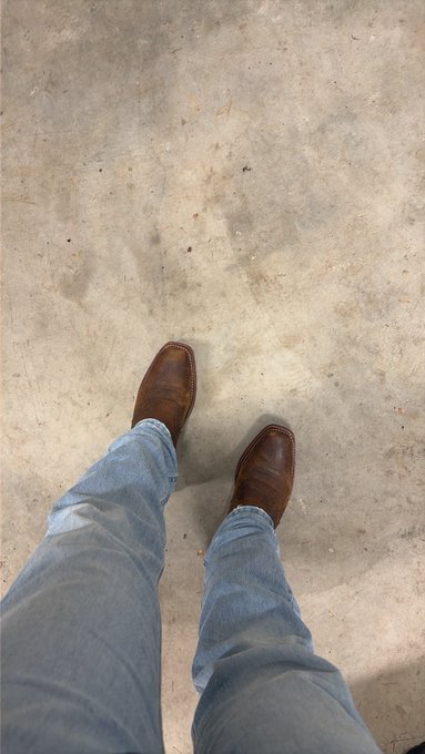 A person's perspective looking down at their brown leather cowboy boots and blue jeans standing on a concrete floor.