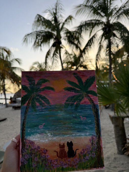 A hand holds up a small painting depicting a tropical sunset scene with palm trees, flamingos, and two cats sitting on a beach, with real palm trees in the background.