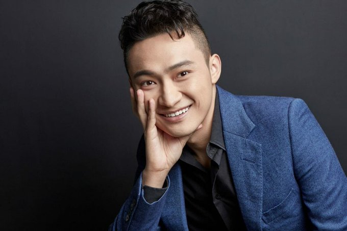 A professional studio portrait of a smiling East Asian man wearing a navy blue blazer over a dark shirt, posing with his hand against his cheek against a dark gray background.