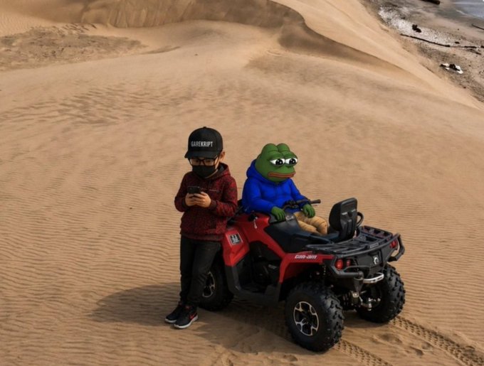 A child wearing a black cap and maroon hoodie stands next to a red Can-Am ATV with a Pepe the Frog figure seated on it, set against a sandy desert landscape.