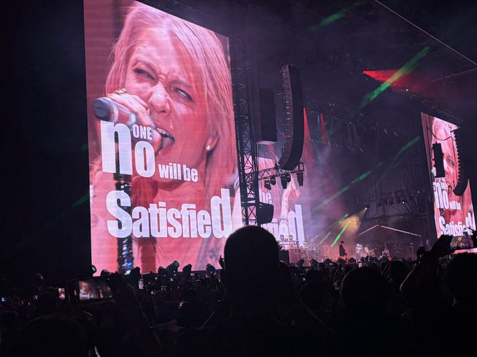 A concert scene shows a singer performing on a large screen displaying the lyrics 'no one will be satisfied,' with green laser lights illuminating the venue and a crowd watching below.