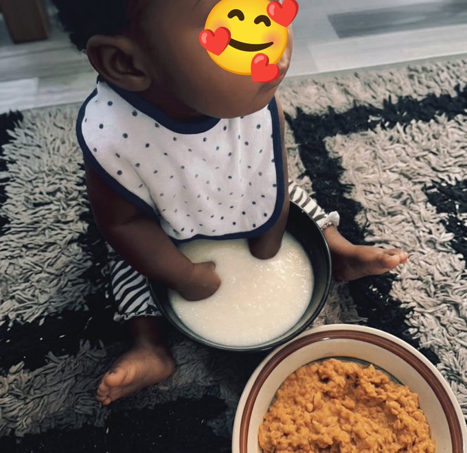 A photo of a baby wearing a polka-dot bib sitting on a shag rug with a bowl of white food (likely porridge or pap) between their legs and a bowl of orange stew beside them, with a heart-eyes emoji covering the baby's face.
