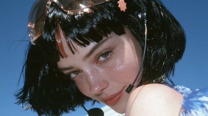 A close-up portrait of a young woman with a black bob haircut wearing reflective orange-tinted sunglasses and a headset microphone, set against a blue background.