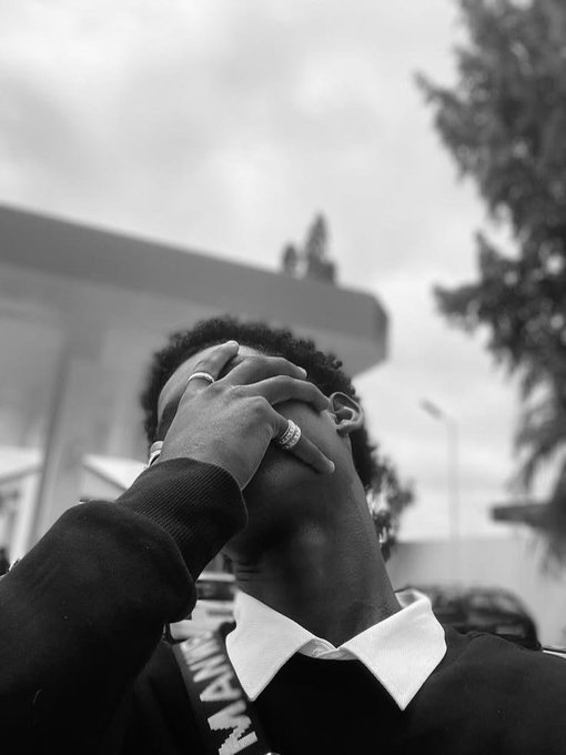 A black-and-white photograph of a young person covering their face with both hands, wearing rings and a lanyard, taken from a low angle against a cloudy sky.