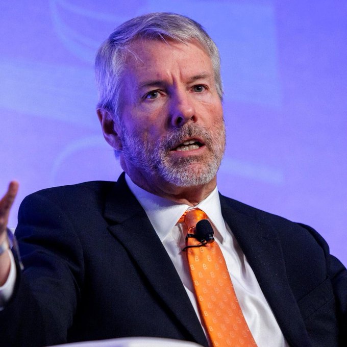 The image features a man wearing an orange tie, sitting down with his hands outstretched as if he's speaking or gesturing. The background of the image is blue, which contrasts with the man's attire. The caption below the image reads "MICHAEL SAYLOR: 'WE'S BUYING Bitcoin SO FAR THIS YEAR... THAT INSTRUMENT ALONE GOES TO buy 2-3x THE B bitcoin supply produced by miners." This suggests that Michael Saylors is talking about a significant amount of Bitcoin being bought, possibly in the millions or even billions. The image also includes a microphone and a handbag, which further supports the idea that this could be an event related to cryptocurrency or a public speaking engagement on the subject.