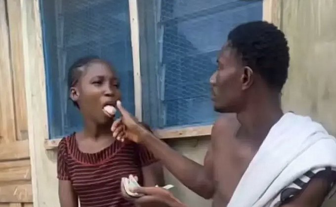 A young man in a white towel applies something to a young woman's mouth with his finger in what appears to be an outdoor scene near a building.