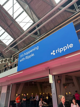 The image shows a group of people gathered around a blue sign that says "RIPPLE" above a restaurant or cafe. The sign appears to be advertising something related to cryptocurrency, possibly a new cryptocurrency exchange or an event related to the crypto world. There are several people in the scene, some standing and others sitting at tables inside the establishment. A few individuals can also be seen walking around outside of the building.