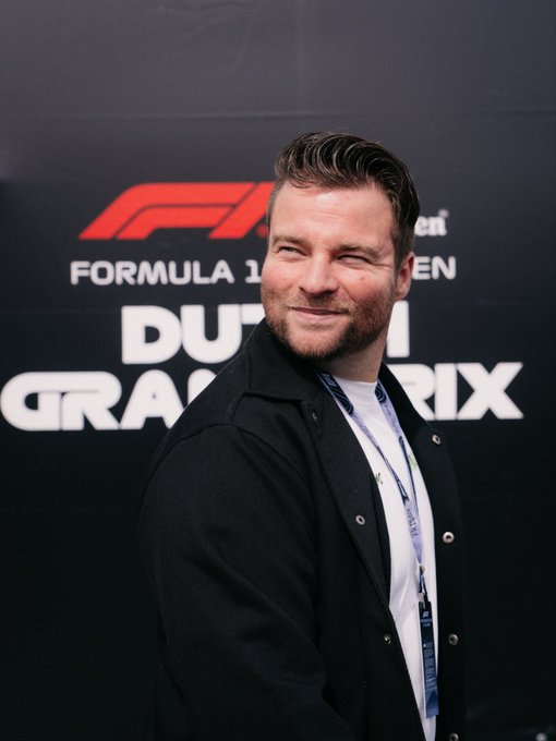 A man with short brown hair and a beard smiles at the camera while standing in front of a backdrop displaying the Formula 1 logo and "DUBAI GRAND PRIX" text, though the caption references the #DutchGP at Zandvoort with sponsorship from @cryptocom.