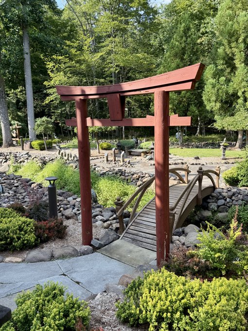 A traditional red Japanese torii gate stands in a tranquil Japanese garden, framed by a stone bridge and lush greenery under bright sunlight.
