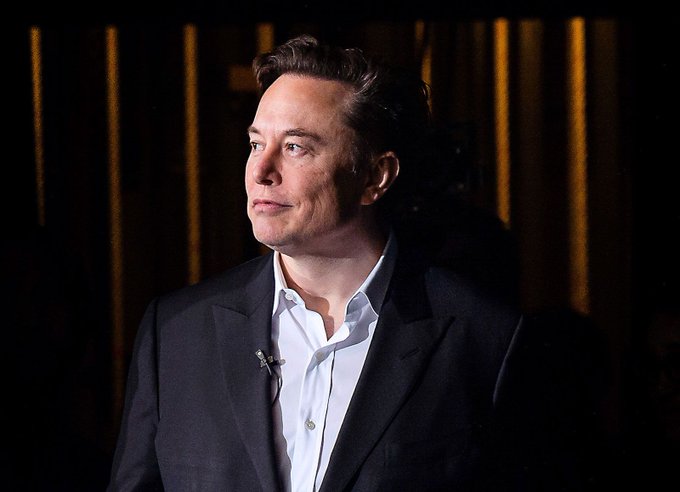 A man in a dark suit and white shirt looks off to the side during what appears to be an interview or speaking engagement, with a small lavalier microphone clipped to his lapel. The image likely accompanies the post referencing Elon Musk, whose name appears in the caption alongside mentions of @X and @SpaceX, suggesting the pictured individual is Musk discussing stablecoin payouts for creators on the X platform.