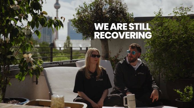 A man and woman wearing sunglasses sit on a rooftop patio with the text 'WE ARE STILL RECOVERING' overlaid on the image.