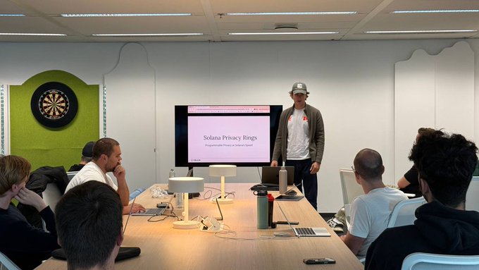 A presenter wearing a cap stands in front of a screen displaying 'Solana Privacy Rings', speaking to an audience seated around a conference table in a room with a dartboard.
