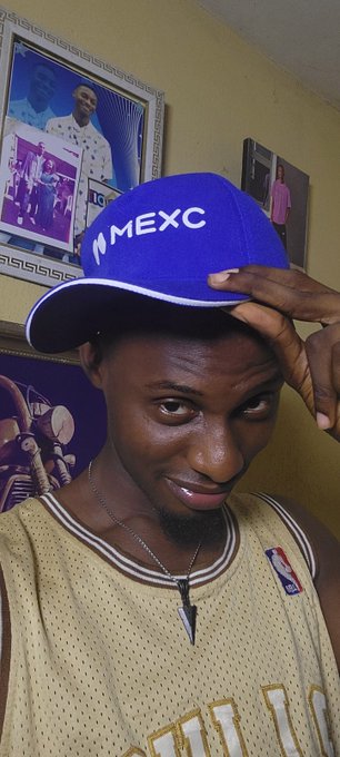 A man wearing a blue "MEXC" cap and a beige NBA jersey poses indoors, with framed photos on the yellow wall behind him. The image relates to the post's dismissive tone toward crypto/NFT promotion by showing someone visibly wearing a MEXC (cryptocurrency exchange) branded hat.