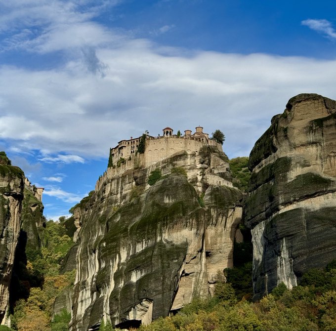 A monastery perched atop a towering sandstone rock formation with surrounding cliffs and lush greenery under a partly cloudy blue sky, characteristic of the Meteora monasteries in Greece.