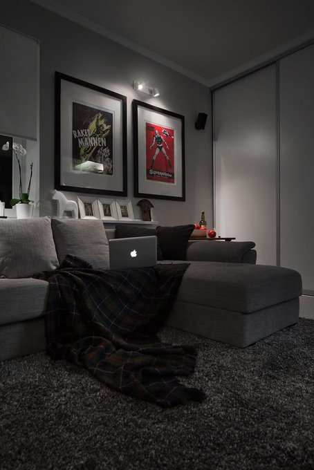 A dimly lit modern living room with a gray sectional sofa, plaid throw blanket, and MacBook, featuring framed posters including 'Bake Köket' and what appears to be a Schwarzenegger image on the wall.