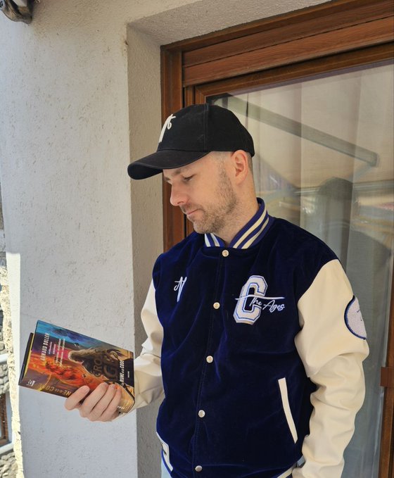 A man wearing a navy and white varsity jacket and a black 'NY' cap looks down at a box he is holding, appearing to read the text 'LEAGEND BARES' and 'Furyc' on its cover.