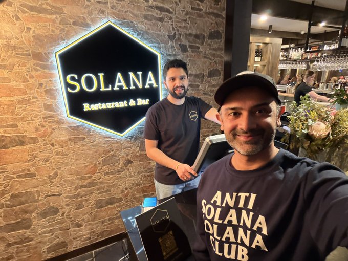 Two men pose in front of a glowing neon 'SOLANA Restaurant & Bar' sign; one wears a shirt reading 'ANTI SOLANA SOLANA CLUB'.
