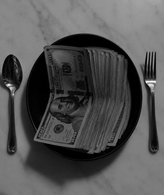 A black-and-white image of a black plate filled with stacks of $100 US bills, with a spoon on the left and a fork on the right, sitting on a marble surface.