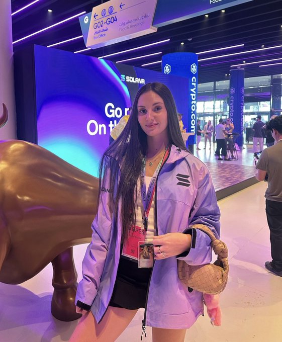 A woman wearing a lavender jacket and a VIP pass poses in front of a 'CRYPTO.COM' sign and a large golden bull statue at an event.