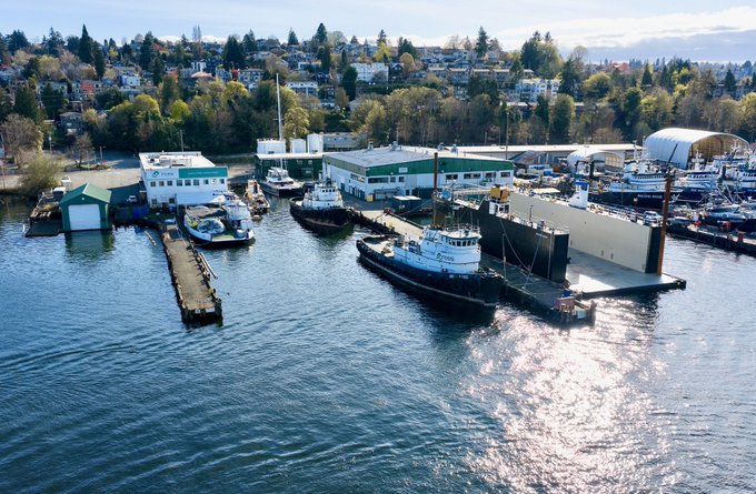 Aerial view of a harbor with tugboats and ships docked at piers, with a residential hillside neighborhood in the background.