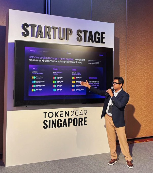 A man in a navy blazer and tan pants presents at the 'STARTUP STAGE' for 'TOKEN2049 SINGAPORE', pointing to a screen about 'Balsore scales through meme capital'.