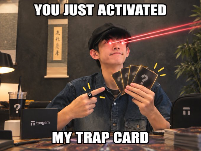 A man in a denim shirt and black cap winks as red laser beams shoot from his eyes while holding trading cards with question marks, with text 'YOU JUST ACTIVATED MY TRAP CARD' and a 'tangem' branded device on the desk.
