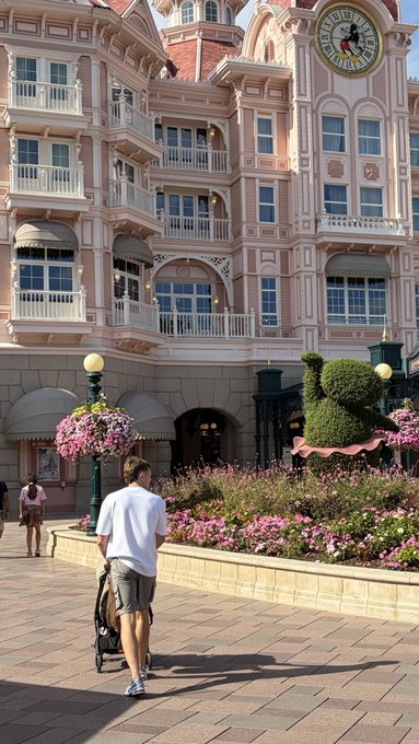 A man in a white shirt pushes a stroller past the iconic pink facade of Disney's Boardwalk Resort, where a Mickey Mouse topiary and flower baskets line the brick pathway. The image captures the "Disney dad" moment from the post, showing him enjoying a sunny day at the park with his child.