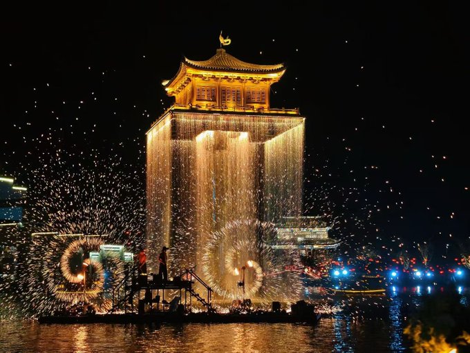 A nighttime scene featuring a glowing traditional Chinese pavilion with cascading lights, accompanied by performers creating large circular fire-spinning displays on a raft in the water below.