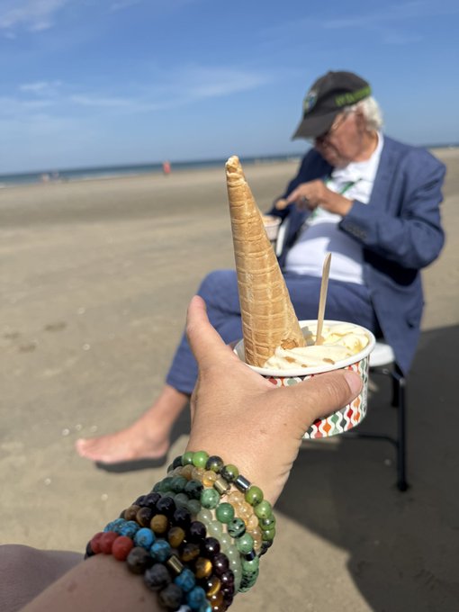 A first-person view of a hand with beaded bracelets holding an empty ice cream cone on a beach, with a man in a blue suit sitting in a chair in the background.
