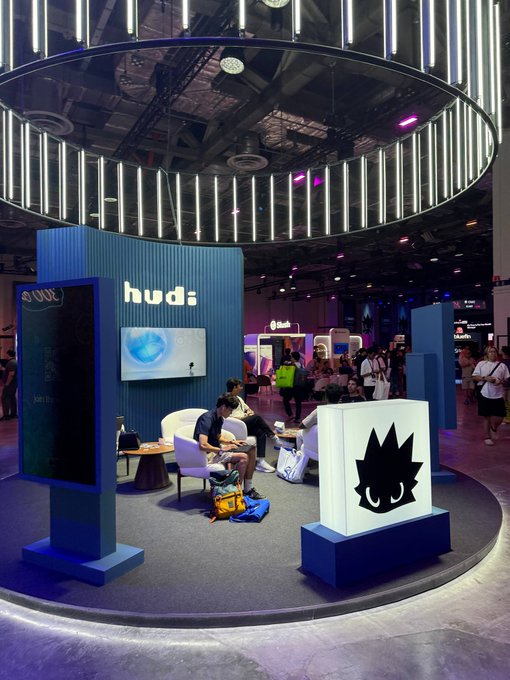 A convention or expo booth for 'hud' featuring a large illuminated circular ceiling display, a screen showing a face, and people sitting in a lounge area.
