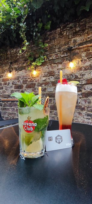 Two cocktails on a dark table at the Havana Club bar, including a mojito-style drink and a tall layered sunset drink, set against a brick wall with string lights and hanging plants.