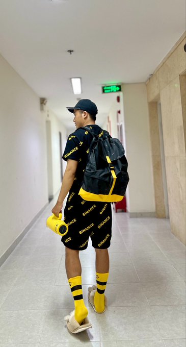 A person walks down a hallway wearing a black t-shirt, shorts, and a backpack all branded with the "BINANCE" logo, paired with yellow socks and holding a yellow camera. The post in Chinese translates to "May I ask you to sign this outfit?" — tagging @binance and @cz_binance (Binance founder Changpeng Zhao), suggesting the outfit is themed after the Binance brand and the wearer is playfully requesting CZ's autograph.