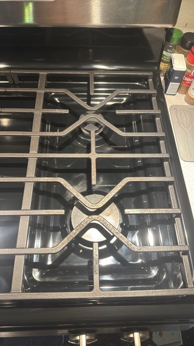 A close-up view of a gas stove featuring cast iron grates with an X-shaped cross pattern, with a bottle of "Simply" seasoning visible on the counter to the right, humorously illustrating the post's joke about "XRP burner grates" playing on the cryptocurrency ticker.