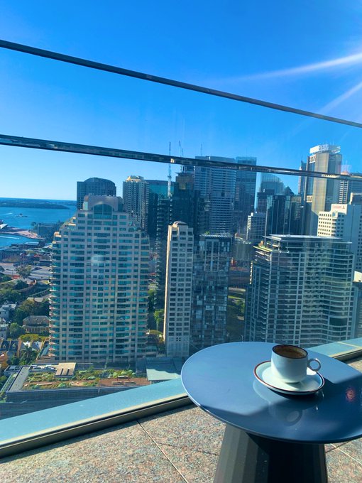 A coffee cup sits on a small table by the window of a high-rise building, overlooking a sprawling cityscape with modern skyscrapers and a waterfront under a clear blue sky. The serene morning view perfectly matches the Arabic greeting "صباح الخيرات" (Good morning, full of blessings), offering a peaceful start to the day.