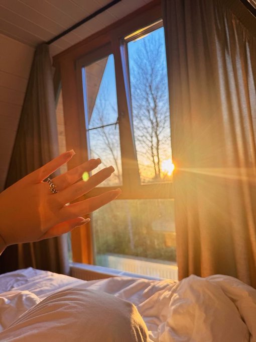 A hand reaches toward a window where golden sunset light streams through, illuminating a bedroom with white bedding and tan curtains.