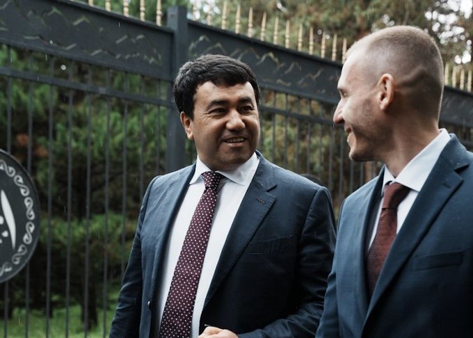 Two men in business suits standing and conversing in front of a black wrought iron gate with greenery in the background.