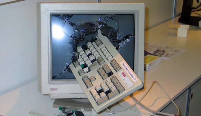 A damaged Compaq CRT monitor with a shattered screen, with a beige computer keyboard partially lodged into the broken glass.