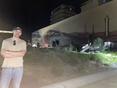 The image features a man standing next to a building, with his arms crossed over his chest. He appears to be posing for a photo or enjoying the view of the city at night. The building behind him has an interesting design that resembles a starbase, which is indicated by its unique shape and the presence of stars on it. The post accompanying this image reads "Happy Starbase Day!". This suggests that the man might have taken the photo to celebrate the occasion or simply appreciate the artistic aspect of the building's design.