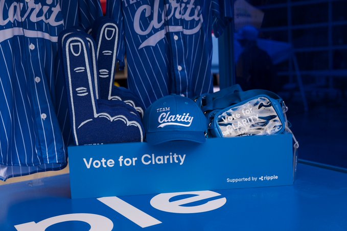 The image features a blue box with white text that reads "Vote for Clarity" and "Team Clarity", along with a baseball cap and other items related to the theme of clarity, such as a sign or poster. The context suggests that this is likely an advertisement or promotional material for a political campaign or event focused on promoting clarity in decision-making and understanding complex issues.