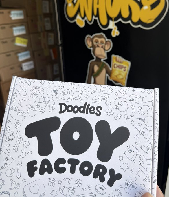 A hand holding a white box labeled 'Doodles TOY FACTORY' with doodle illustrations, in front of what appears to be a shop with a cartoon monkey mascot and storage boxes.