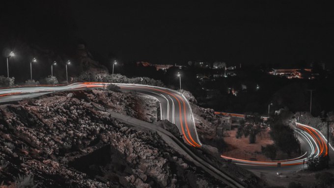 A long-exposure nighttime photograph captures sweeping light trails of red and white from vehicles navigating a winding mountain road, illuminated by scattered streetlamps.