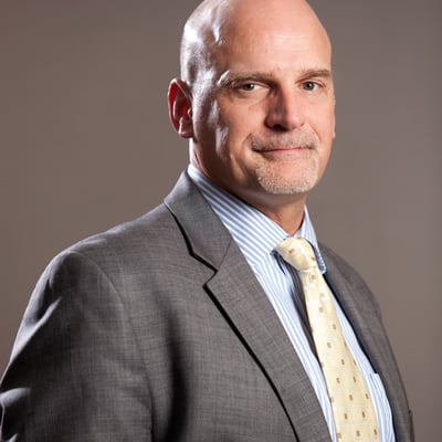 The image shows a bald man with a gray goatee wearing a gray suit, blue striped shirt, and a cream/yellow patterned tie, posed in a professional portrait against a gray background—likely a stock photo used to illustrate the post about Mike supposedly having "Bitcoin Derangement Syndrome."