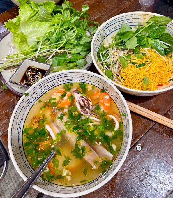 A bowl of Vietnamese soup (likely bún riêu or bún bò) garnished with herbs, alongside a plate of fresh greens, bean sprouts, and yellow noodles, with chopsticks and a small dish of dipping sauce on a wooden table.