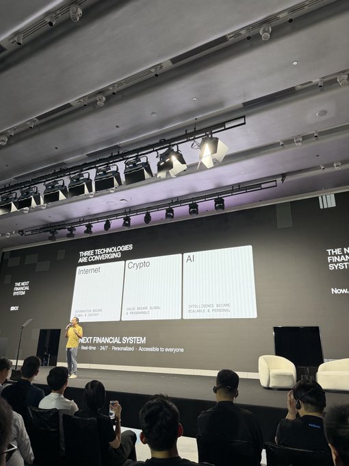 A man in a yellow shirt stands on a stage giving a presentation to an audience. The large screen behind him is titled 'THREE TECHNOLOGIES ARE CONVERGING' and lists 'Internet', 'Crypto', and 'AI', along with the text 'NEXT FINANCIAL SYSTEM'.
