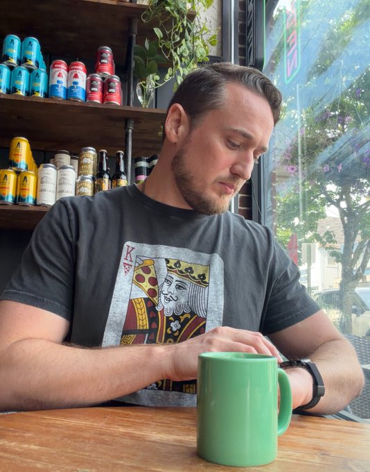 A man wearing a black King of Hearts poker card t-shirt sitting at a wooden table with a green mug, with a window and beverage cans visible in the background.