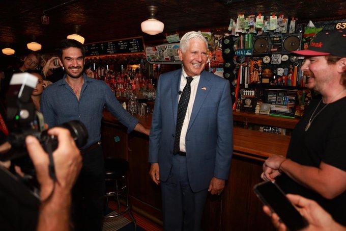 A photograph of an older man in a blue suit smiling in a bar setting, surrounded by younger men and photographers with cameras.