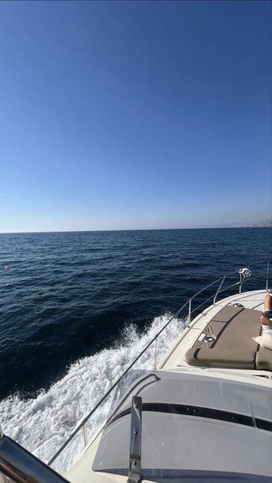 A view from the bow of a moving boat cutting through the blue ocean under a clear sky, with a person's legs visible as they sit on the deck.