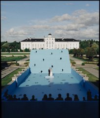 A scene showing a grand white palace in the background while the foreground features a surreal blue landscape with a small figure standing on what appears to be a large blue surface, observed by silhouetted onlookers.