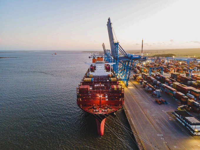 An aerial view of a large cargo ship docked at a port at sunset, with massive shipping cranes and stacks of containers visible alongside the vessel.