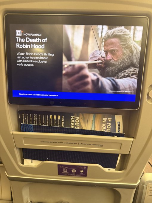 A United Airlines seatback touchscreen displays 'The Death of Robin Hood' as 'Now Playing' for exclusive early access, with magazines stored in the seat pocket below.
